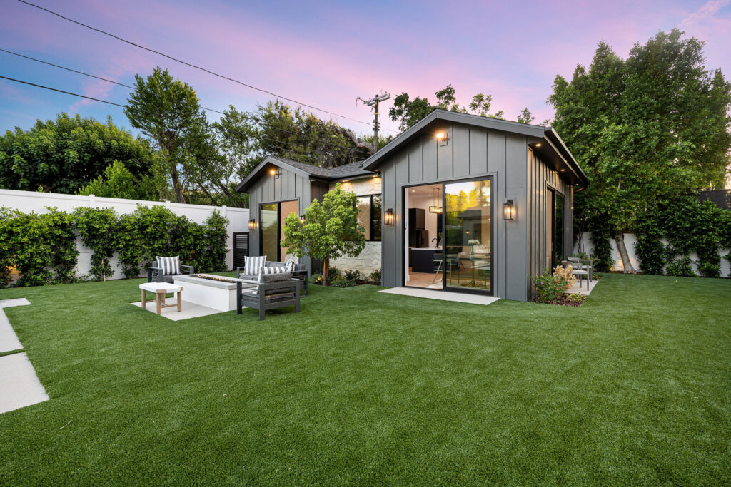 Modern backyard with a small house, patio furniture, and lush green lawn at sunset.
