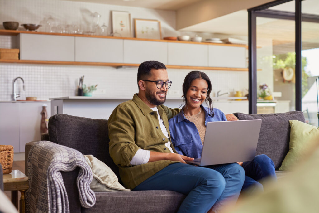 Young mixed race couple watching movie on laptop while relaxing on a weekend at home. Smiling indian man and beautiful woman sitting on sofa while using laptop. Happy mid adult caual man and beautiful hispanic woman sitting on couch while surfing the net.